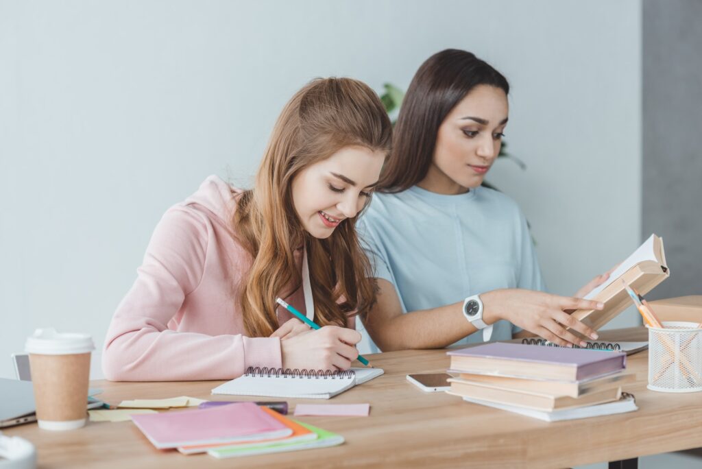 multiethnic girls studying together and writing in copybook and reading book at table