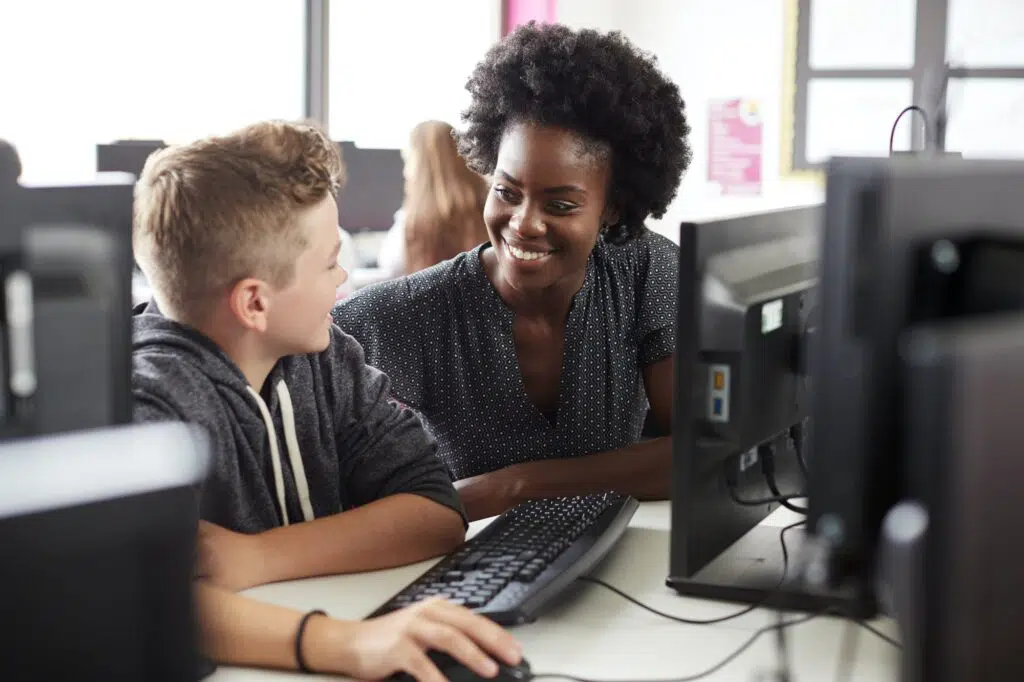 Teacher Helping Male High School Student Working at Screen In Computer Class